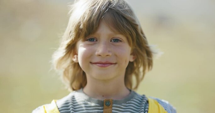 Charming smiling 5-6 years old outside. Portrait of child with long hair. Blond-haired and blue-eyed boy looks into frame. Dimples on cheeks, yellow jacket. Grace and calm, meditative mood. 