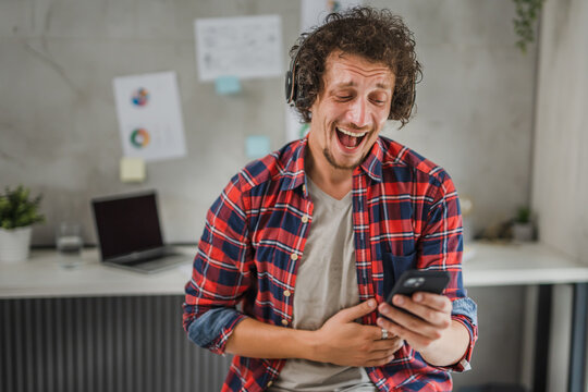 One young man with headphones laughing and looking at mobile phone, making video calll, taking photos selfies, modern lifestyle concept 