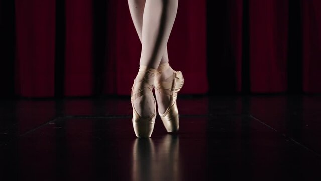 In a slow and close-up shot, a ballet dancer twirls on the tips of her toes in a circle. Only her legs from the knees down are visible. She dances on a theater stage.
