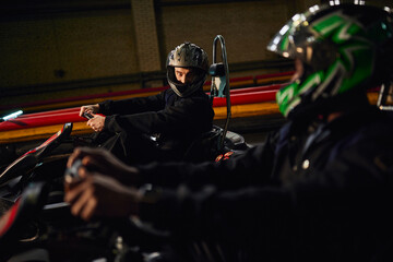 go kart driver in helmet looking at african american friend and  driving on circuit, competition