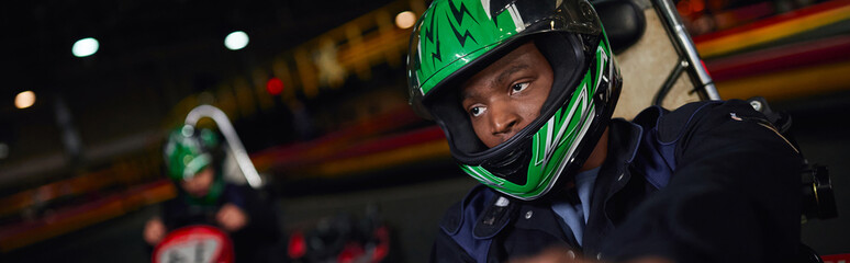 african american man in helmet driving go kart on circuit near friend on blurred backdrop, banner