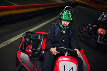 focused man in sportswear and helmet driving sport car for karting near friend on indoor circuit