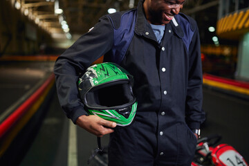 cheerful african american man in sportswear holding helmet and standing in karting track circuit