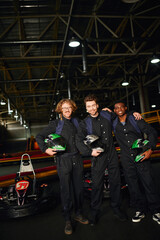 group of multicultural and happy racers in protective suits hugging and holding helmets near go kart