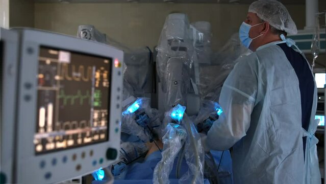 Medical equipment and monitors in background. Robotic surgery in sterile environment. Surgeon controls robot with multiple arms and performs surgical procedure.