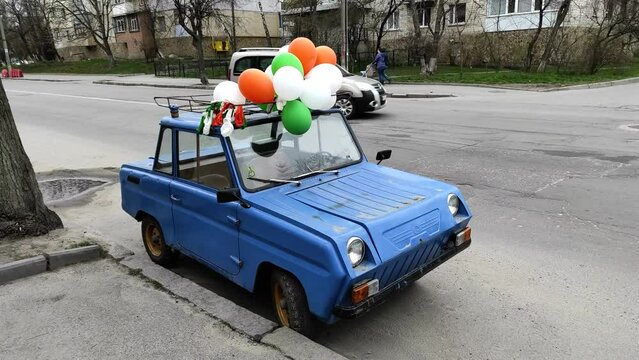 A vintage small Soviet car with balloons on the roof stands in the roadway.