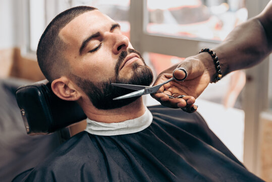 Black barber cutting beard of client with scissors