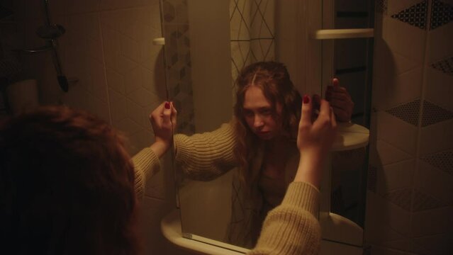 A tired sleepy young caucasian girl with long curly hair and a beige jacket looking in bathroom wall mirror with tungsten light. The problem of insomnia and work fatigue. View of the reflection
