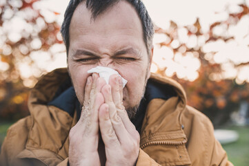 A bearded man in  stylish jacket blows his nose into a paper napkin outdoors in an autumn park....