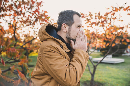 A bearded man in  stylish jacket blows his nose into a paper napkin outdoors in an autumn park. Seasonal autumn cold