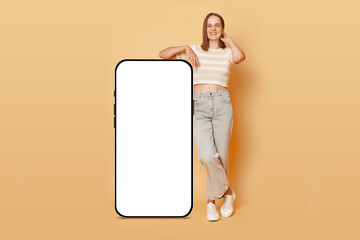 Full length portrait of smiling satisfied woman standing near big mobile cell phone with blank screen workspace area isolated on beige background space for promotional text.