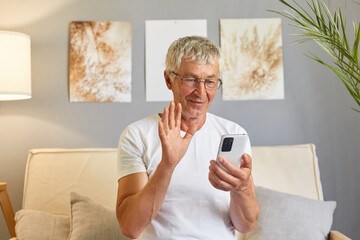 Joyful happy older cellphone user man wearing white T-shirt waving hand hello at gadget frontal camera, speaking on video call, online conference chat, talking to family, resting on home couch.