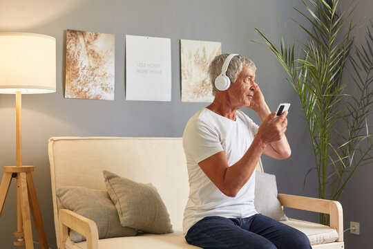 Side view of senior gentleman dressed in T-shirt sitting on couch, holding his mobile phone and headphones, listening to music or podcast lookign away at window attentively listening to audio book