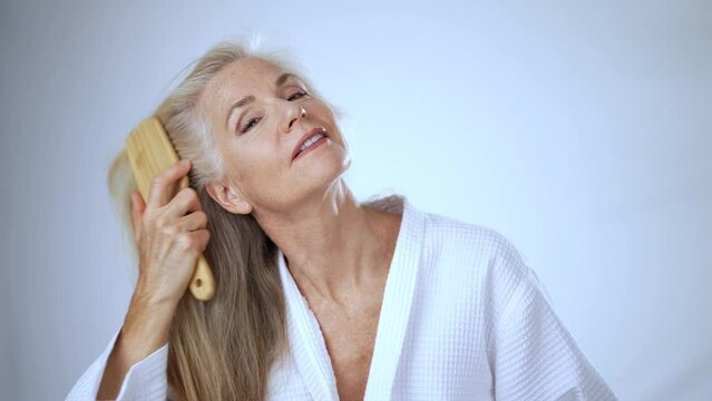 Portrait of middle-aged caucasian old mature woman in bathrobe combing clean hair with hairbrush in home bathroom beautiful senior lady self care beauty routine natural shampoo balm conditioner