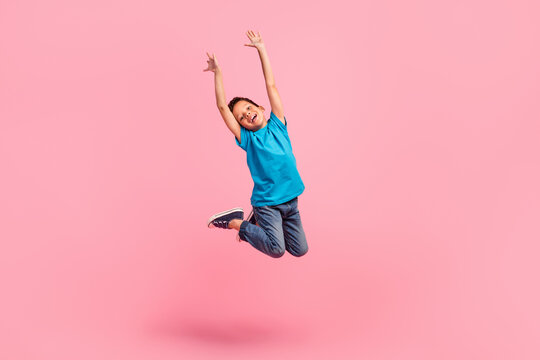 Full length photo of impressed cheerful small boy wear blue t-shirt rising hands arms jumping high isolated pink color background