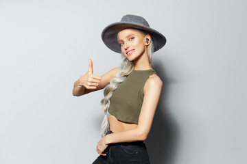 Studio portrait of attractive happy blonde girl with wireless earbuds in ears, showing thumb up, on white background. Wearing grey hat and green shirt.
