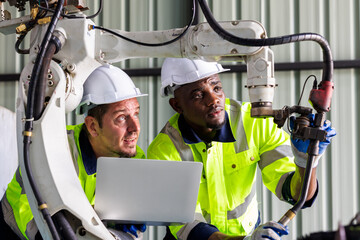 Supporting Engineer team, african american and caucasian man use laptop to control and testing Artificial Intelligent welding robotic automatic arm machine in modern factory. industry 4.0 Technology.