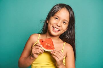 Happy ethnic girl child standing with piece of watermelon against turquoise backdrop