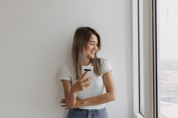 Fototapeta premium Asian woman holding cellphone standing look at window.