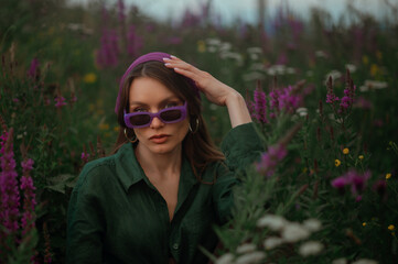 Fashionable confident woman wearing trendy purple rectangular sunglasses, bandana, hoop earrings, green linen shirt, sitting, posing in summer field. Copy, empty space for text