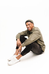 Vertical photo of serene happy african-american guy in hat sitting on the floor in relaxed pose isolated on white background, positive man with attractive smile looking at camera
