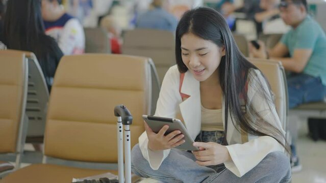 Attractive asian young woman traveler talking on video coll to friend or family through tablet while waiting for her flight sitting on seat in departure gate at airport terminal.