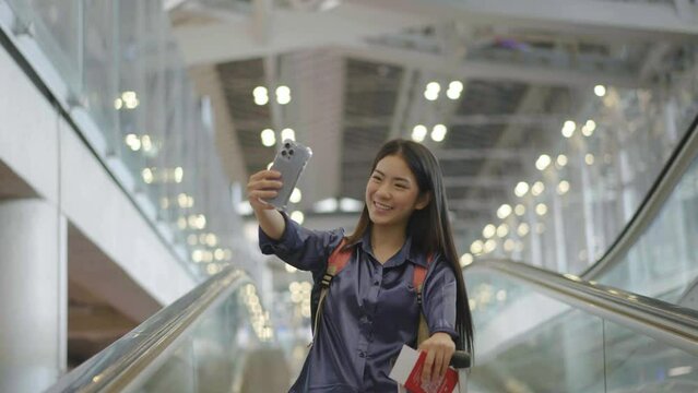 Young asian woman standing on escalator and making selfie using smartphone camera or video call at the airport, Smiling female talking in video conference by smartphone