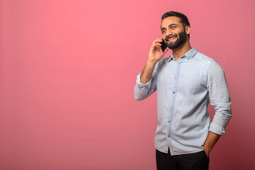 Cheerful Indian male entrepreneur in casual jeans shirt is talking on the smartphone, smiling...