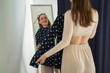 Pretty young woman standing and choosing clothes, trying on a shirt, a woman getting dressed, getting ready before leaving for work, looking at her reflection in the mirror.