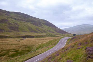 Fototapeta na wymiar Roads turning in Scotland highlands scenery. Road leaving to infinity among mountains, peaks with heather in full bloom. Summer photo with dramatic clouds, on the road, roadtrip, travelling, visiting