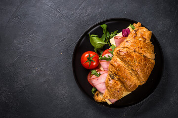 Croissant sandwich with ham, cheese and vegetables on wooden cutting board with cup of coffeee at black stone table. Top view with copy space.