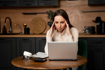 A young brunette woman is stressed using a laptop while sitting in the kitchen.