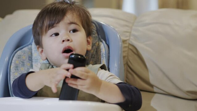 Cute Asian baby sits in a feeding chair, holds a remote control in his hands