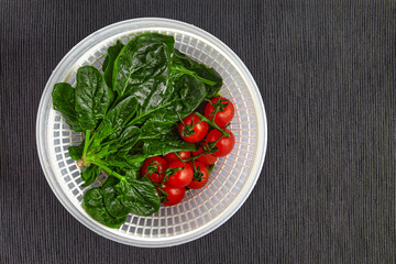 Washed green spinach leaves and cherry tomatoes on a branch in a plastic colander on a black textile tablecloth top view with copy space