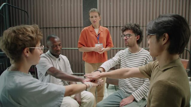 Young multi-ethnic people sitting in circle with eyes closed and stacking hands together while female psychologist telling affirmation on group therapy session