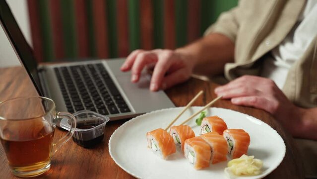 Close-up of a young handsome man dressed in a beige shirt scrolling the social media feed on a laptop, eating sushi with salmon