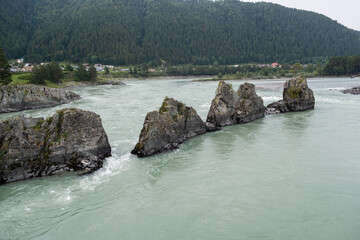 A wide, full-flowing mountain river with a fast current. Large stones stick out of the water. The...