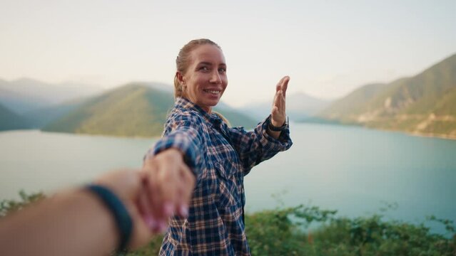 Follow me video with woman calling man waving hand gesture holding his hand looking at amazing mountains river nature view at sunset. Smiling happy female tourist on vacation. Travel, tourism concept.