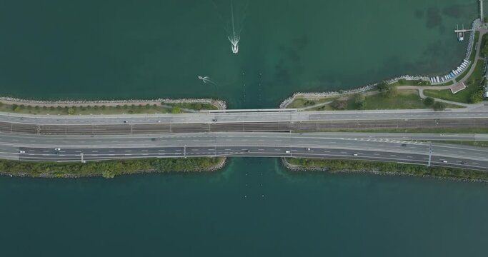 Aerial view of vehicles driving on a road crossing the Lugano lake with a bridge and a boat sailing under the bridge, Melide, Ticino, Switzerland.