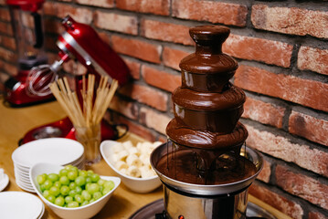 Chocolate fondue fountain in a pot next to a red brick wall. Catering table.