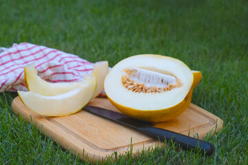 a cut melon lies on a wooden board next to it lies a black knife and a towel in a red cage on a green lawn