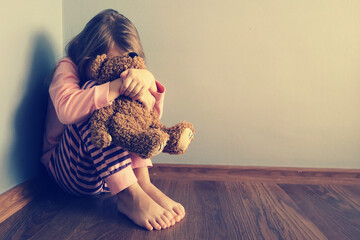 The child is sitting sad scared on the floor in the corner and hugs a toy. Little girl crying with...
