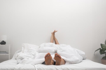 Female with bare feet put out from under cover lies on comfortable bed in light colors suffering from insomnia in lit bedroom of housing disease with lack of sleep in adulthood