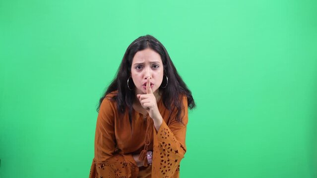 Beautiful Young indian woman casually dressed seriously showing silence gesture on camera over removable Chroma green background. Don't tell expression