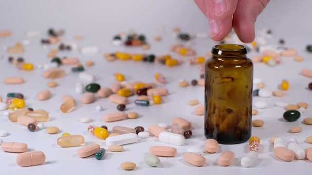 Caucasian male hand putting colourful pills and capsules into a traditional brown pharmacy bottle. Treatment and prevention of diseases. Assortment of medicinal pharmaceutical products. 4K