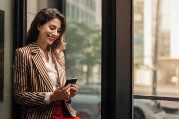Fototapeta premium A woman in a corporate office space laughs while looking at her phone, radiating pride and confidence. She's dressed in unique, eye-catching attire that adds flair to the professional setting.