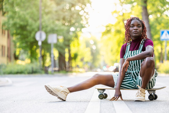 Portrait of a black non-binary person with a skateboard sitting on the street
 - Powered by Adobe