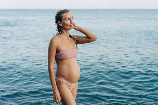 A young pregnant woman stands on the seashore after swimming.
