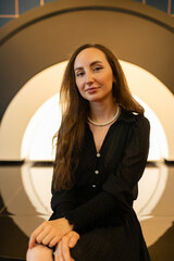 Female poses for photo shoot in studio with minimalistic decorations. Brunette woman with hands crossed on knees looking at camera