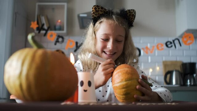 Happy little girl preparing for Halloween party, painting face on pumpkin in the kitchen 
